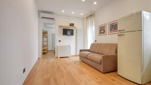 Un espacio habitable pequeño con piso de madera, sofá color beige, refrigerador blanco, televisor montado en la pared y un radiador.