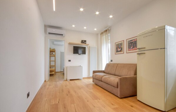 Un espace de vie compact doté d’un plancher en bois, d’un canapé beige, d’un réfrigérateur blanc, d’un téléviseur fixé au mur et d’un radiateur.