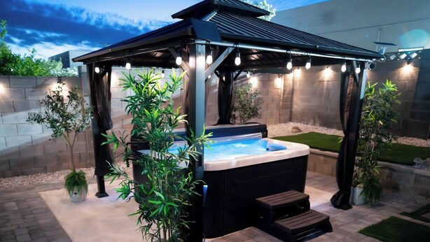 Un jacuzzi bajo un porche con luces de cuerda, rodeado de plantas en macetas y un patio de ladrillo.