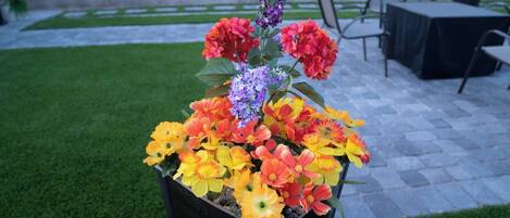 A flower arrangement in a planter on a patio with a stone floor and chairs in the background.