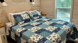 A bedroom with a wooden wall, a bed with a blue and white quilt, two wall-mounted lamps, and a window with a view of greenery.