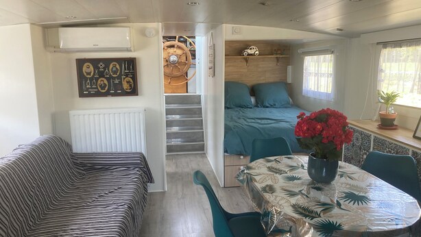 A compact room with a bed, a sofa, a dining table with chairs, a radiator, a wooden headboard, and a window with curtains. - Péniche Marie-lou (Thourotte)