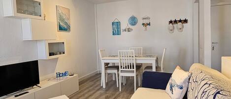 Ein Wohnzimmer mit Fernseher, weißen Möbeln, einem Essbereich mit Stühlen und einem gemusterten Sofa.