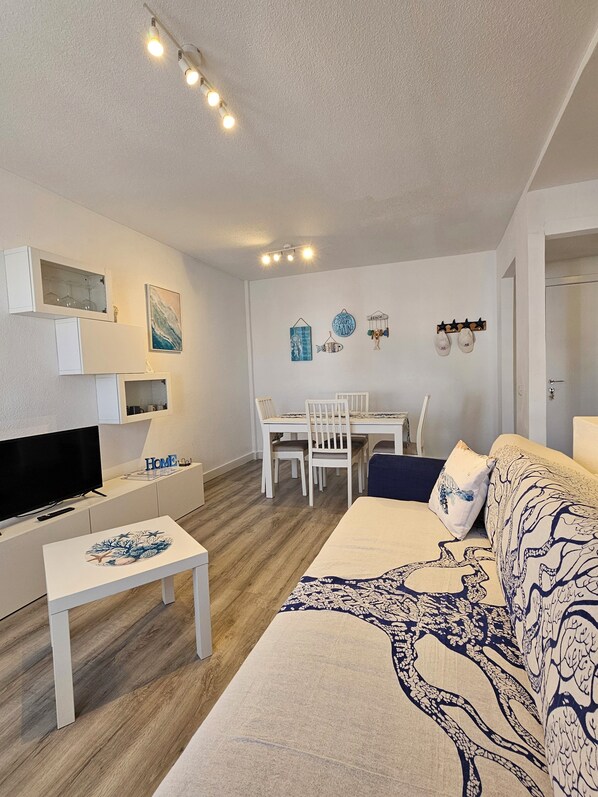Una sala de estar con televisor, muebles blancos, un comedor con sillas y un sofá estampado.