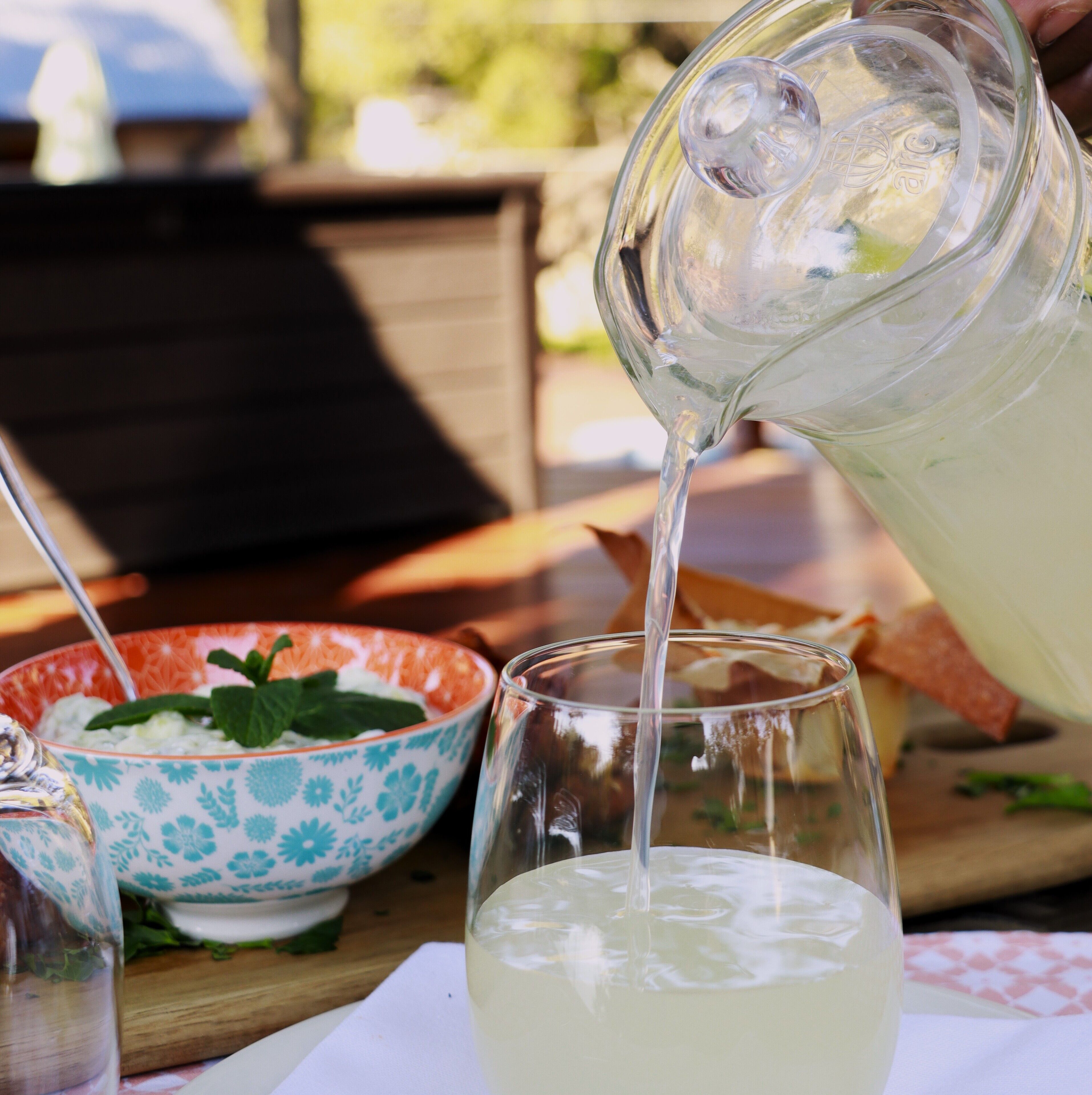 A pitcher pouring a clear liquid into a glass, with a bowl of food and a bottle in the background.