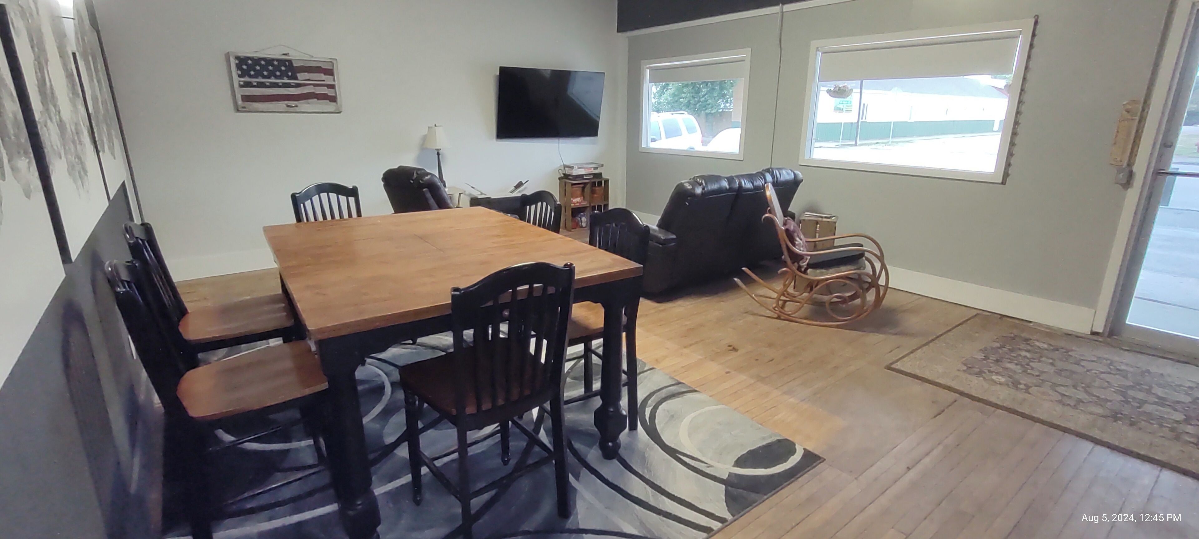 A dining area with a wooden table and chairs, a television mounted on the wall, and two windows with a view of the outside.