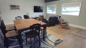 A dining area with a wooden table and chairs, a television mounted on the wall, and two windows with a view of the outside.