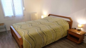 A bedroom with a wooden bed, a bedside table with a lamp, and a radiator on the wall. - Maison de Vacances Pour 2 Personnes. Animal Accepté - Plage-mer 10 min (Grues)