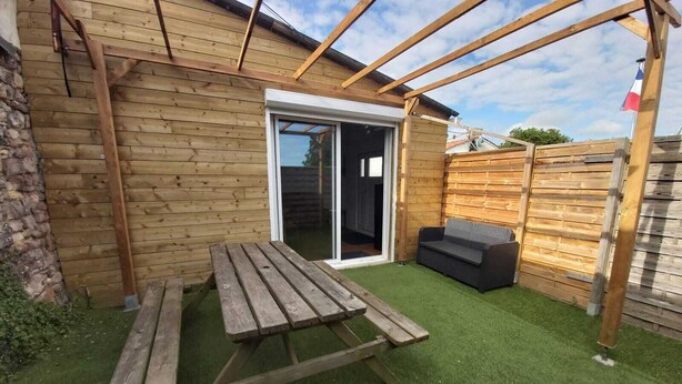 A wooden deck with a picnic table and a sofa, a sliding glass door, and a French flag. - Maison de Vacances Pour 2 Personnes. Animal Accepté - Plage-mer 10 min (Grues)