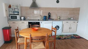 A compact kitchen with a round wooden table, four chairs, a microwave, a washing machine, and a sink. - Maison de Vacances Pour 2 Personnes. Animal Accepté - Plage-mer 10 min (Grues)