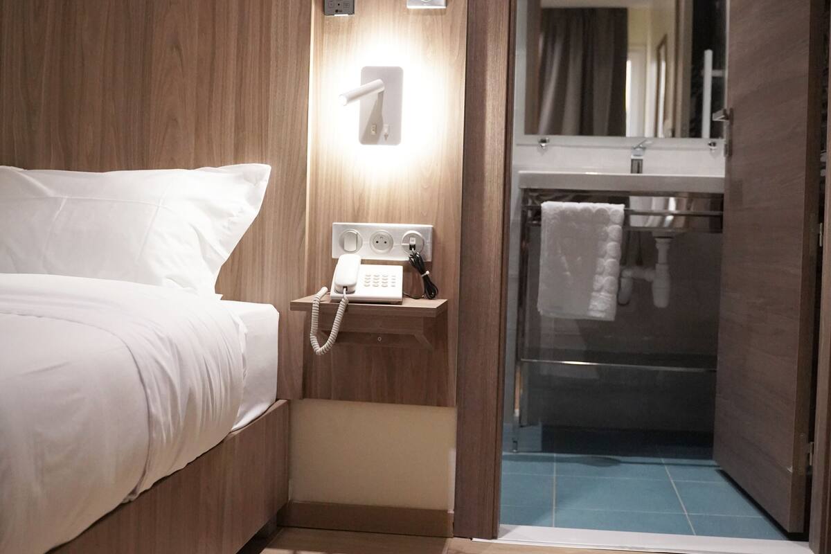 Quarto de hotel com cabeceira de madeira, telefone ao lado da cama e porta de vidro do chuveiro.
