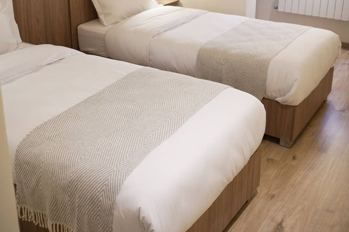 Quarto de hotel com duas camas, cabeceiras de madeira, mesa de cabeceira, luminária e janela com vista.