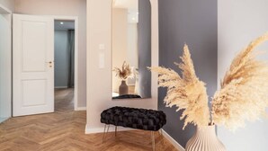 A hallway with a white door, a black cushioned bench, a tall mirror, and pampas grass in a vase. - NEW LUXUS Suite | TWO Kingbeds | Netflix | Center (Augsburg)