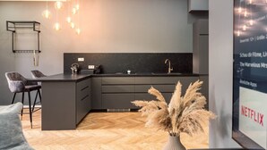 A modern kitchen with a black countertop, grey cabinets, and a wooden floor. There are two bar stools and a vase with dried plants. - NEW LUXUS Suite | TWO Kingbeds | Netflix | Center (Augsburg)