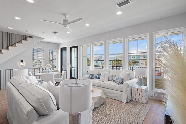 A modern living room with a staircase, large windows, and a ceiling fan.