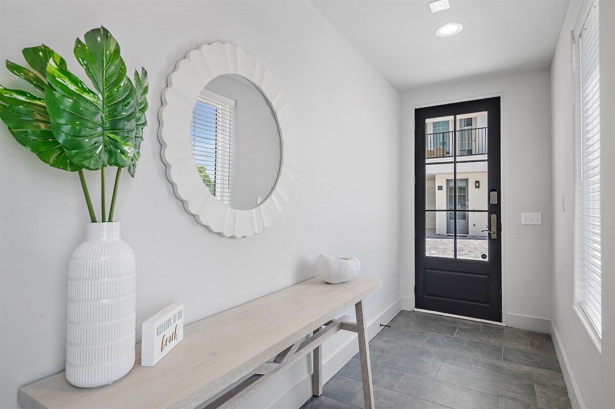 A hallway with a white door, a round mirror, a white vase with green leaves, and a bench.