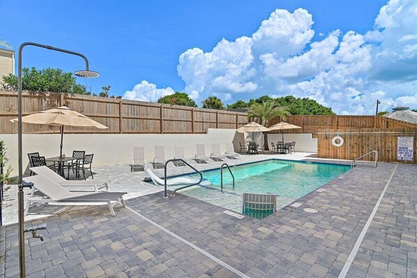 A swimming pool with lounge chairs and umbrellas, surrounded by a wooden fence and a brick patio.