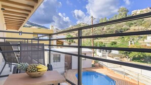 A balcony with a pool, a table with a bowl of fruit, and a view of a hillside with buildings. - Your Family Beach Getaway: Pool, BBQ & Ocean at Your Doorstep! (Ligaria)
