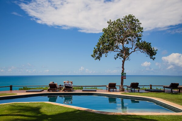 A pool with lounge chairs and a tree, overlooking a clear blue ocean. - Beijo do Vento Pousada (Porto Seguro)