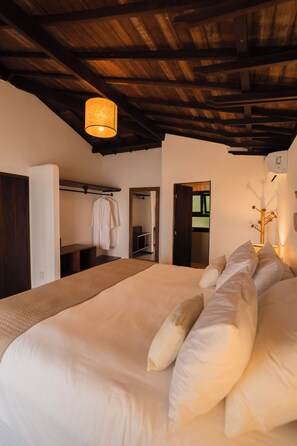 A bedroom with a large bed, wooden ceiling beams, a hanging light fixture, a coat rack, and a door leading to another room. - Beijo do Vento Pousada (Porto Seguro)
