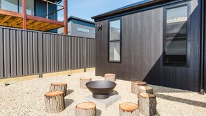 A modern outdoor seating area with a fire pit, surrounded by wooden stumps, in front of a building with large windows and a metal fence. - 16B Rawson Street - Brand new Build (Jindabyne)