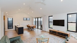 A spacious living area with a dining table, a kitchen island, a flat-screen TV, and a large window offering a view of the outdoors. - 16B Rawson Street - Brand new Build (Jindabyne)