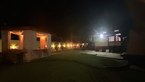 Una vista nocturna de un patio con un pequeño edificio, una ventana iluminada y un camino bien iluminado.