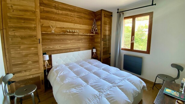 A bedroom with a wooden headboard, a bed with white bedding, a bedside table with a lamp, a chair, and a window with a view of greenery. - Cozy T2 Apartment for 3 in Formiguères - Close to Shops and Ski Slopes (Formiguères)