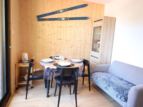 A compact dining area with a wooden wall, a table set with plates, glasses, and cutlery, and a grey sofa with a patterned cover. - Downtown Bagnères-de-Luchon 1BR with balcony, WiFi, pets allowed (Bagnères-de-Luchon)