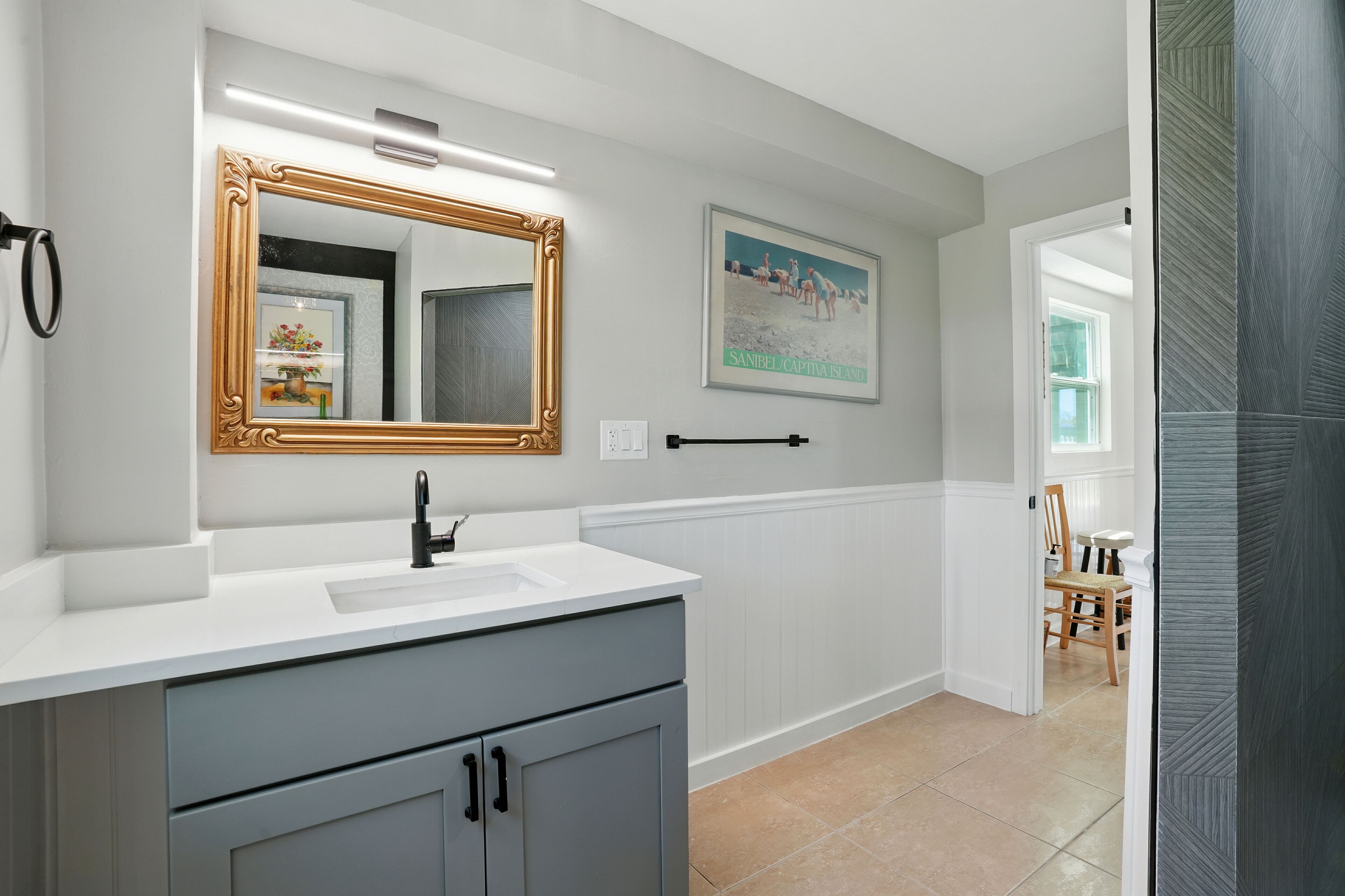 A bathroom with a white vanity, a gold-framed mirror, and a painting of a beach scene.