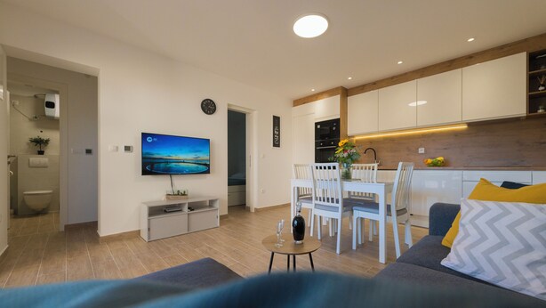A modern living room with a sofa, a dining table with chairs, a TV mounted on the wall, and a kitchen area with cabinets and a microwave. - Apartment with Free Covered Parking (Zadar)