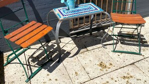 Un espace patio avec une table et deux chaises en bois, un arrosoir bleu et une fenêtre avec des barreaux.