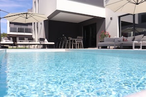 Ett modernt utomhusområde med pool, solstolar och parasoller, ett matbord och en soffa.