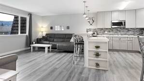Un appartement moderne avec un canapé gris, une table basse blanche et une cuisine dotée de meubles blancs et d’un four à micro-ondes encastré.