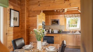 Une cabane en bois avec un coin repas comprenant une table dressée pour deux personnes, un vase avec des fleurs et une cuisine en arrière-plan.