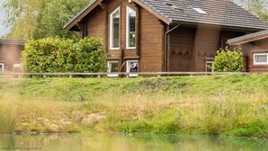 Une cabane en bois dotée d’une grande fenêtre et d’une porte, entourée d’une végétation luxuriante et d’un petit étang.