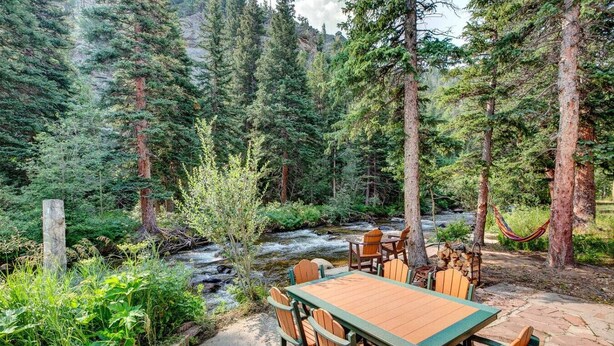 An outdoor dining area with a table and chairs, surrounded by a forest and a stream. - Riverside`s Hideout Cabin- With Hot Tub! (Lyons)