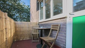 Une petite terrasse extérieure avec une table et des chaises en bois, une porte-fenêtre à treillis et un mur en briques.