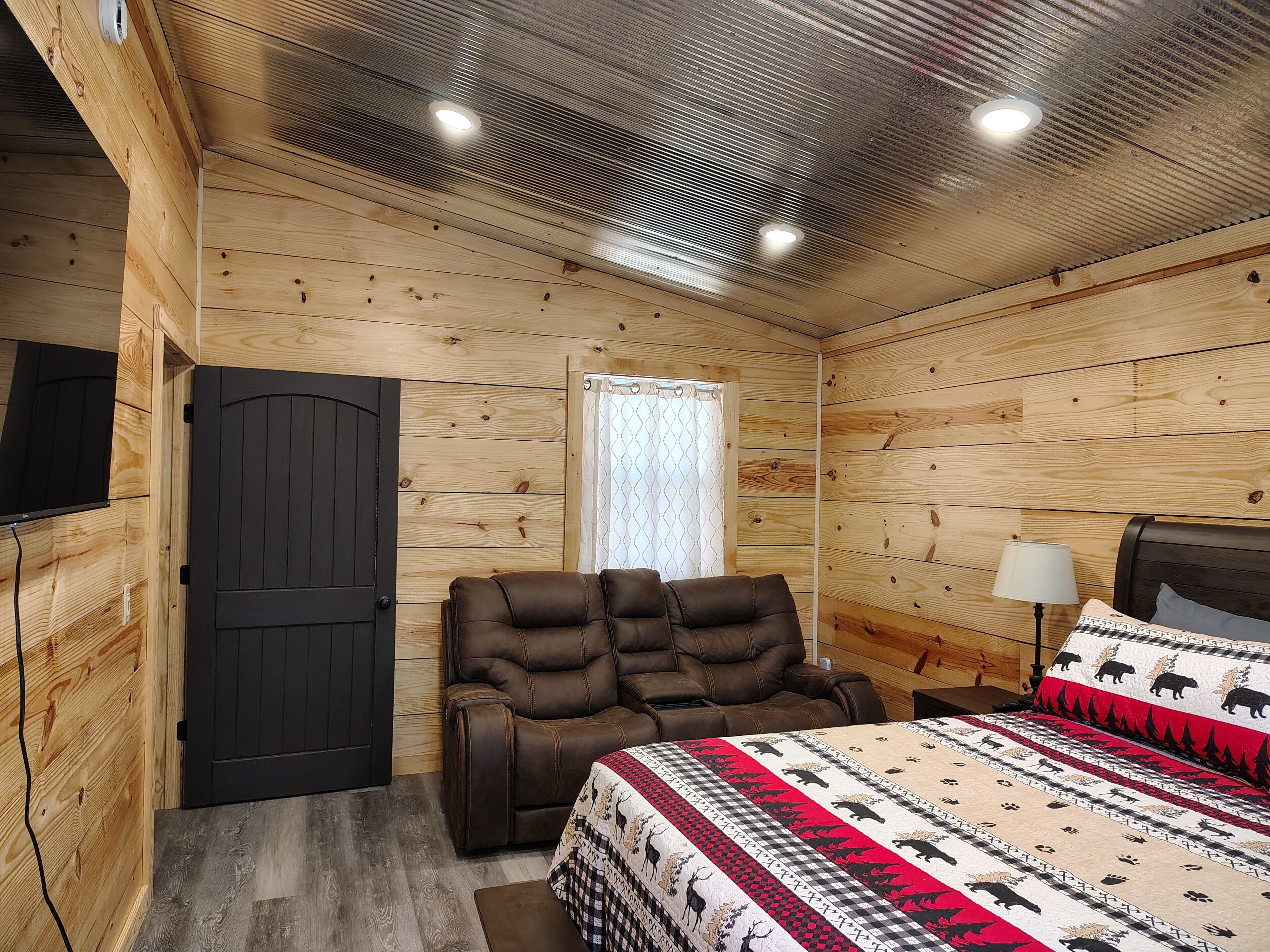 A wooden cabin room with a bed, a recliner, a TV, and a window with curtains.