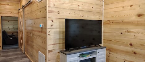 Tunnelmallinen, puinen huone, jossa on suuri, seinälle asennettu taulutelevisio, pieni pöytä, jossa on lehti, ja taustalla näkyy tuoli.