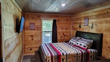A cabin-style bedroom with a wooden ceiling, a bed with patterned bedding, a mounted TV, and a window with a view of trees.