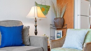 Una sala de estar con un sofá gris, una silla de mimbre, una lámpara, un adorno en la pared y una estantería con un cuadro enmarcado.