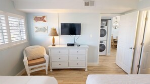 Une chambre avec une commode blanche, un téléviseur, un fauteuil en osier et un lit.
