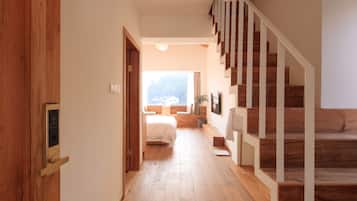 Un pasillo con piso de madera que conduce a un dormitorio con cama y ventana. A la derecha se ve una escalera con escalones de madera y barandillas blancas.