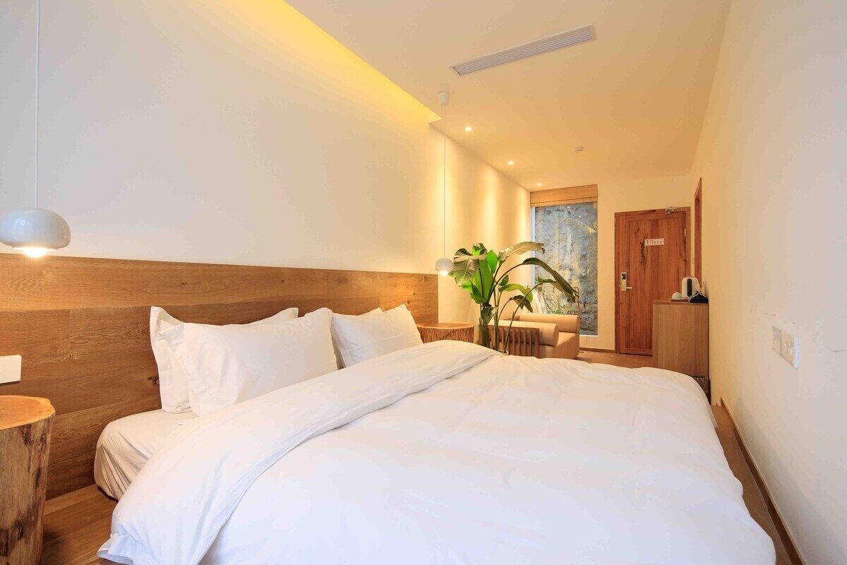 A modern hotel room with a large bed, wooden headboard, bedside tables, and a plant.