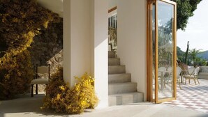 Eine moderne Treppe mit Steinwand und gelbem Laub, die zu einer Glastür führt, welche auf eine Terrasse mit Sitzgelegenheiten im Freien hinausführt.