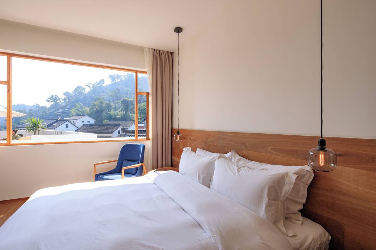 Une chambre d’hôtel avec un grand lit, une tête de lit en bois, une fenêtre donnant sur des maisons et des arbres, et une chaise bleue.