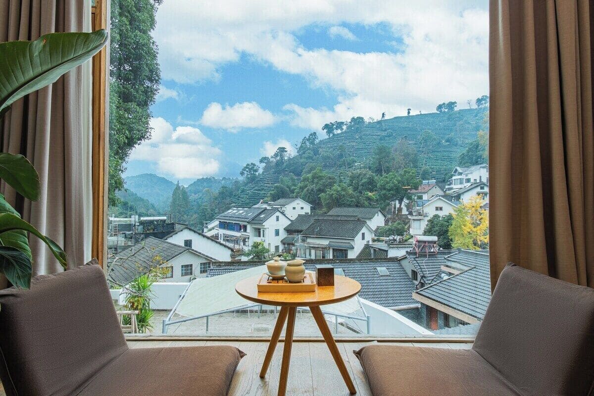 Une pièce avec vue sur des maisons à étages et des collines. On y trouve une table ronde en bois, sur laquelle est posé un plateau et une petite plante.