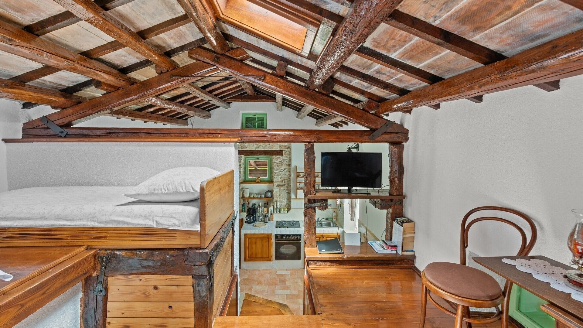 A rustic room with a wooden ceiling, a bed, a television, a small kitchen area, and a chair.