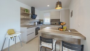 Eine moderne Küche mit Holzausziehtisch, Backofen, Mikrowelle und Kaffeemaschine. Auf der Arbeitsfläche steht ein Hochstuhl für Kinder und ein Obstkorb.